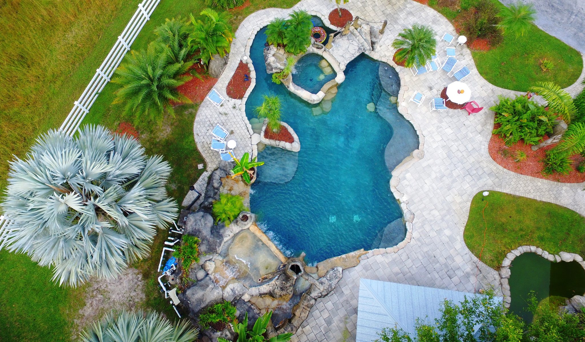 Aerial drone view of the resort-style pool with rock waterfall grotto, hot tub, lounge chairs, putting green, and lush tropical landscaping